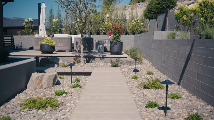 Modern backyard featuring a composite walkway leading to a Techo-Bloc Borealis paver deck with outdoor lounge seating, concrete block retaining walls, decorative rock, boulder accents, hydrangeas, and potted planters in Lake Country, BC, designed by Creative Roots Landscaping