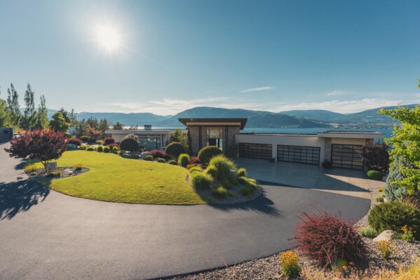 Aerial view of a luxury estate front yard featuring a sweeping circular driveway, manicured lawn island, clipped ball shrubs, ornamental grasses, Japanese maple, and mixed planting beds surrounding a contemporary home with panoramic Okanagan Lake and mountain views, designed by Creative Roots Landscaping in Kelowna, BC