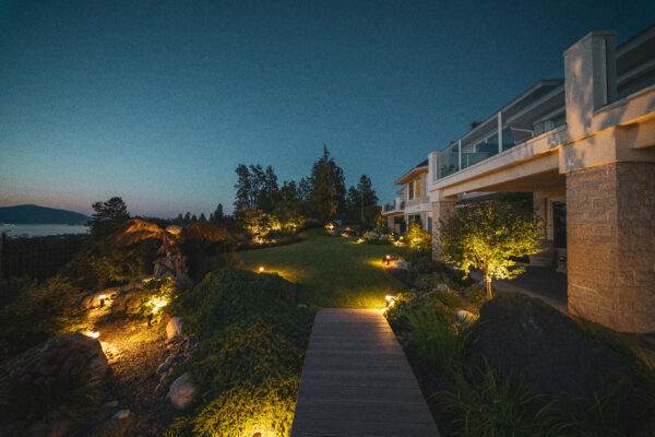 Dusk view of a luxury backyard featuring a composite walkway, professionally lit manicured lawn, uplighted garden beds, boulder accents, a driftwood eagle sculpture, and panoramic Okanagan Lake and mountain views alongside a multi-storey stone home, maintained by Creative Roots Landscaping in Kelowna, BC